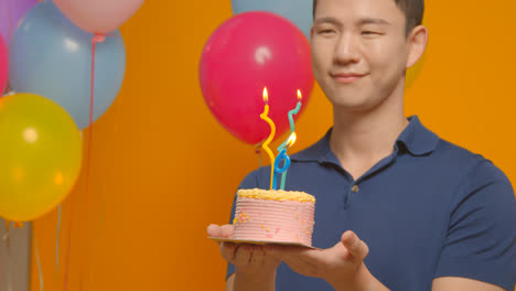 Studio-Portrait-Of-Man-Wearing-Party-Hat-Celebrating-Birthday-Holding-Cake-With-Candles